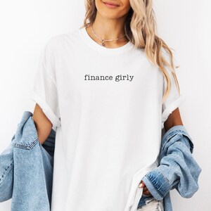 May include: White t-shirt with the text "finance girly" in a black, simple font. The shirt has rolled-up sleeves and is paired with a denim jacket. The overall style is casual and relaxed.