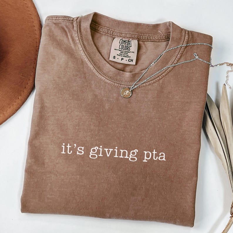 Puede incluir: Una camiseta marr&oacute;n Comfort Colors con la frase "it's giving pta" en letras blancas. Un collar plateado con un peque&ntilde;o colgante descansa sobre la camiseta. Un sombrero marr&oacute;n es parcialmente visible en el fondo.