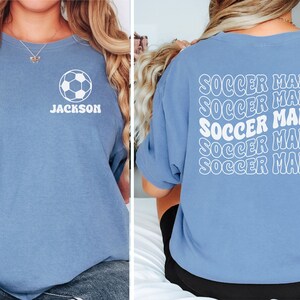 Peut inclure: Un t-shirt bleu clair avec un ballon de football blanc et le nom "JACKSON" sur le devant. Le dos du t-shirt porte la phrase "SOCCER MAMA" r&eacute;p&eacute;t&eacute;e cinq fois dans une police r&eacute;tro.