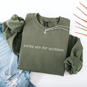 May include: An olive green crewneck sweatshirt featuring the white text "corks are for quitters" printed on the chest. The garment has long sleeves with ribbed cuffs and a soft fleece interior lining, paired with a silver-toned chain necklace.