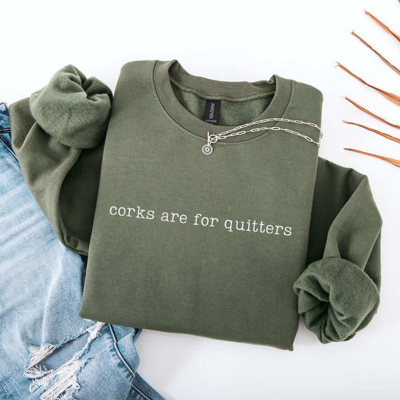 May include: An olive green crewneck sweatshirt featuring the white text "corks are for quitters" printed on the chest. The garment has long sleeves with ribbed cuffs and a soft fleece interior lining, paired with a silver-toned chain necklace.