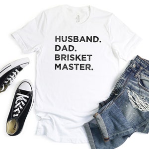 May include: White t-shirt with black text that reads "Husband. Dad. Brisket Master."