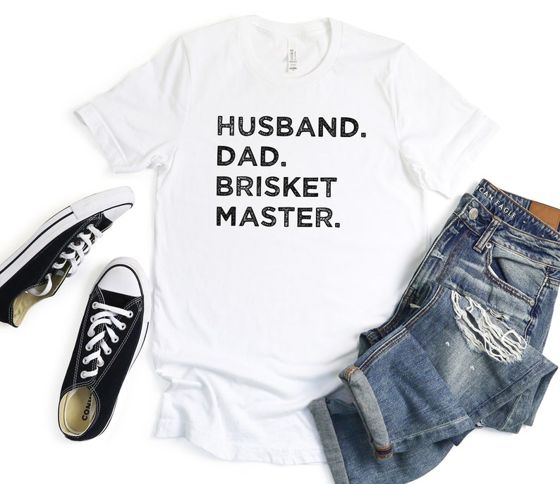 May include: White t-shirt with black text that reads "Husband. Dad. Brisket Master."