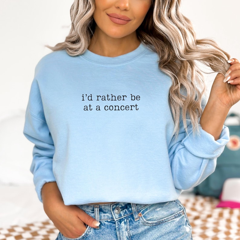 May include: A light blue crewneck sweatshirt featuring the black text "i'd rather be at a concert" printed in a typewriter-style font across the chest. The garment has long sleeves with ribbed cuffs and a ribbed neckline, providing a simple design.