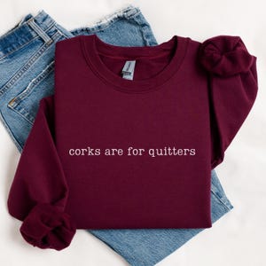 May include: A maroon long-sleeve crewneck sweatshirt featuring the white embroidered text "corks are for quitters" centered on the chest. The garment has a ribbed neckline and cuffs, and is layered over a pair of blue denim jeans.