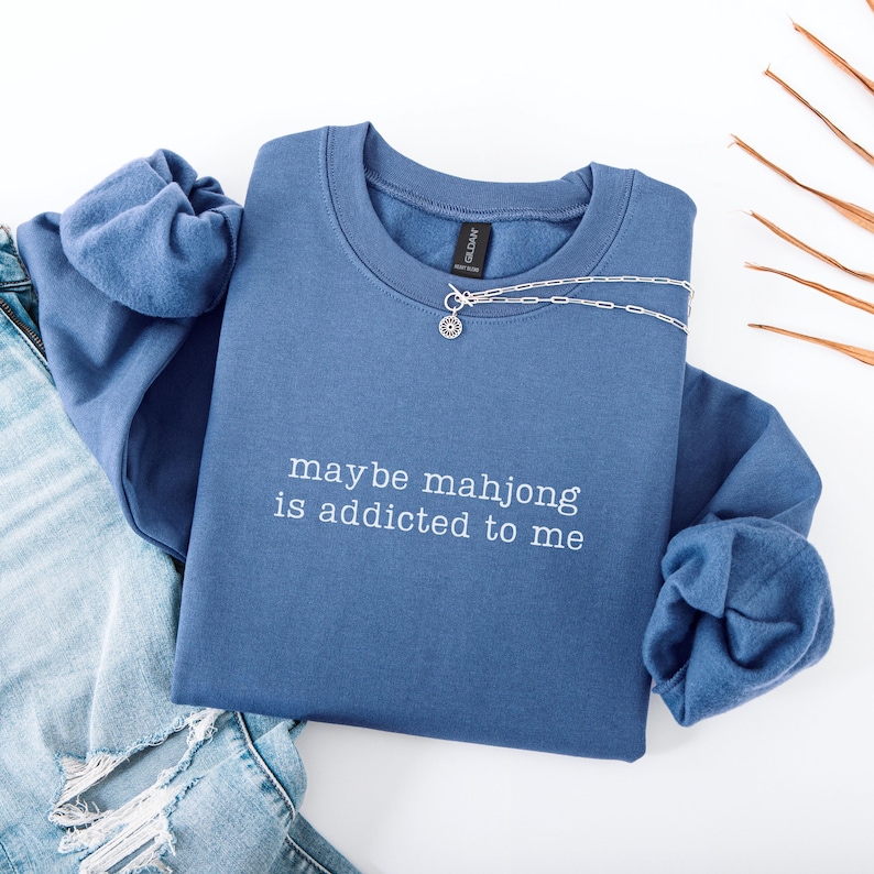 May include: A blue sweatshirt with the phrase "maybe mahjong is addicted to me" in white script. A silver necklace with a circular pendant rests on the sweatshirt. The sweatshirt is paired with blue jeans.