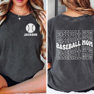 May include: A charcoal gray short-sleeve t-shirt featuring a white baseball graphic with the name JACKSON on the front left chest. The back of the shirt displays the text BASEBALL MOM repeated five times in a wavy, stacked white font design.
