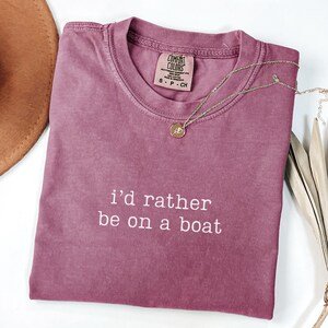 May include: A mauve short-sleeved t-shirt with a crew neckline and the white embroidered text "i'd rather be on a boat" centred on the chest. The shirt features a Comfort Colors brand label inside the collar and a thin gold-toned necklace.