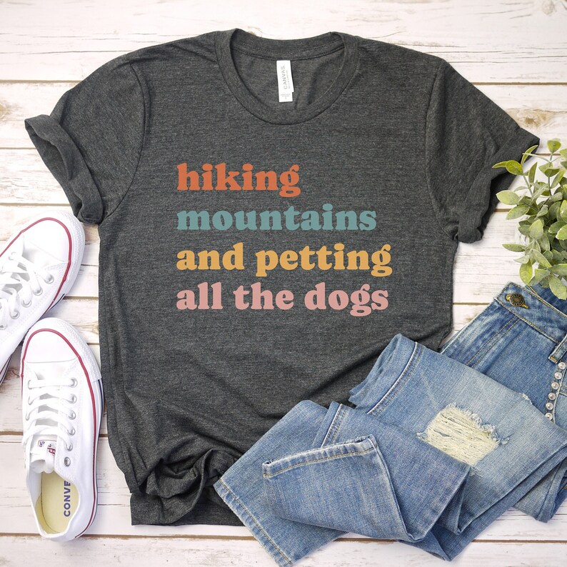 May include: A dark gray t-shirt with the text "hiking mountains and petting all the dogs" in colorful letters. The shirt is laid flat on a white wooden surface with a pair of white sneakers and blue jeans.