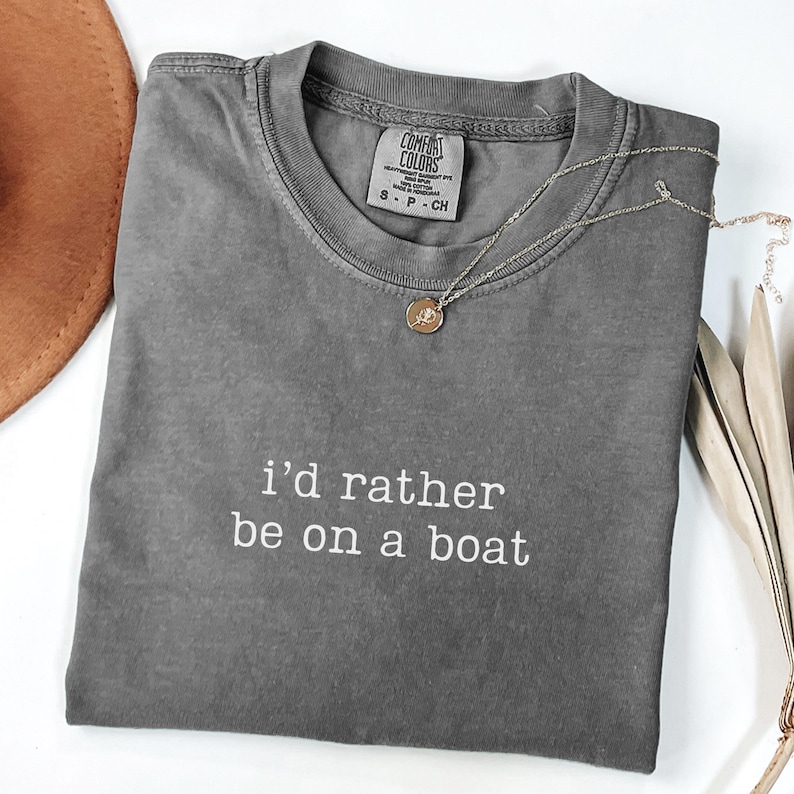 May include: A charcoal grey short-sleeve t-shirt with the white text "i'd rather be on a boat" printed on the chest. The shirt features a ribbed crew neckline and a Comfort Colors brand label inside the collar, accompanied by a thin gold necklace.