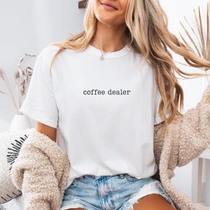 Puede incluir: Una camiseta blanca de manga corta con el texto "coffee dealer" impreso en negro, en min&uacute;sculas y con una tipograf&iacute;a estilo m&aacute;quina de escribir en el centro del pecho. La prenda tiene cuello redondo y se lleva con pantalones cortos de mezclilla.