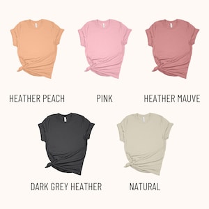 May include: Five different color t-shirts, folded and tied in a knot. The colors are heather peach, pink, heather mauve, dark grey heather, and natural.
