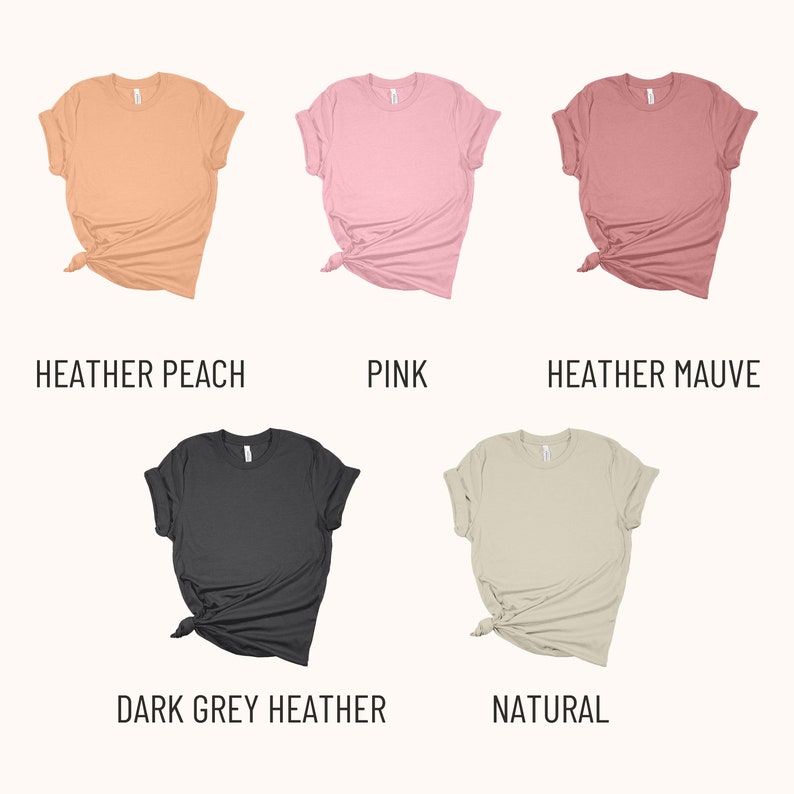 May include: Five different color t-shirts, folded and tied in a knot. The colors are heather peach, pink, heather mauve, dark grey heather, and natural.
