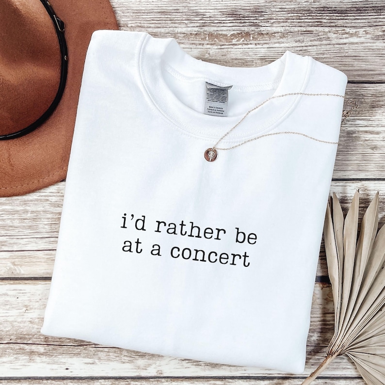 May include: A white crewneck sweatshirt featuring the black text i'd rather be at a concert embroidered on the center chest. The garment has a standard ribbed neckline and a visible interior brand label at the collar, presented in a clean style.