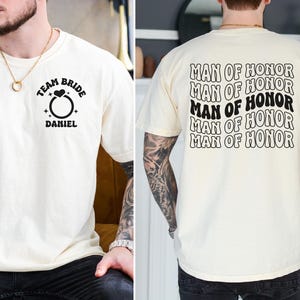 Pode incluir: Camiseta creme com texto e gr&aacute;ficos pretos. A frente apresenta um gr&aacute;fico de anel com as palavras "TEAM BRIDE" acima e "DANIEL" abaixo. A parte de tr&aacute;s da camisa tem as palavras "MAN OF HONOR" repetidas v&aacute;rias vezes.