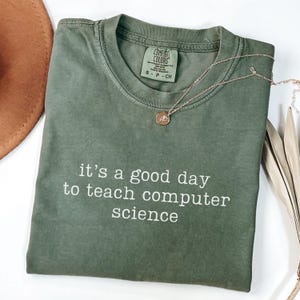 May include: Olive green t-shirt with the text "it's a good day to teach computer science" in white. A gold necklace with a pendant is draped over the shirt. The shirt is folded, and a brown hat is visible in the background.