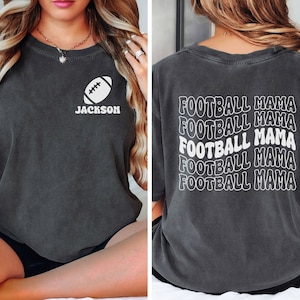 May include: A dark gray t-shirt with a white football graphic and the text "JACKSON" on the front. The back of the shirt has the text "FOOTBALL MAMA" repeated five times in a wavy font.