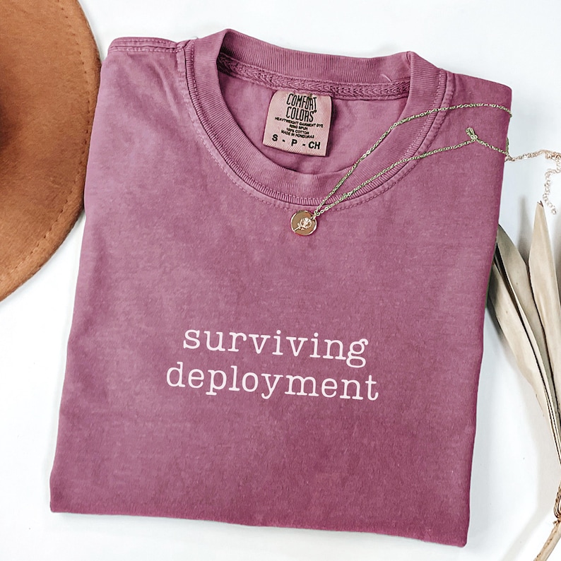 May include: A mauve Comfort Colors t-shirt with the words "surviving deployment" printed in white. A gold-colored necklace with a pendant rests on the shirt. A brown hat is visible in the upper left corner.