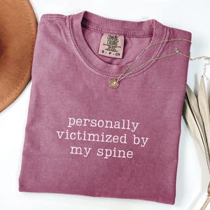 May include: A mauve short-sleeved t-shirt with a crew neckline featuring the white text "personally victimized by my spine" printed on the front. The shirt has a Comfort Colors brand label inside the collar indicating size small, and a gold necklace.
