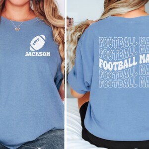May include: A light blue t-shirt with a white football graphic and the name "JACKSON" on the front. The back of the shirt has the phrase "FOOTBALL MAMA" repeated five times in a white, wavy font.