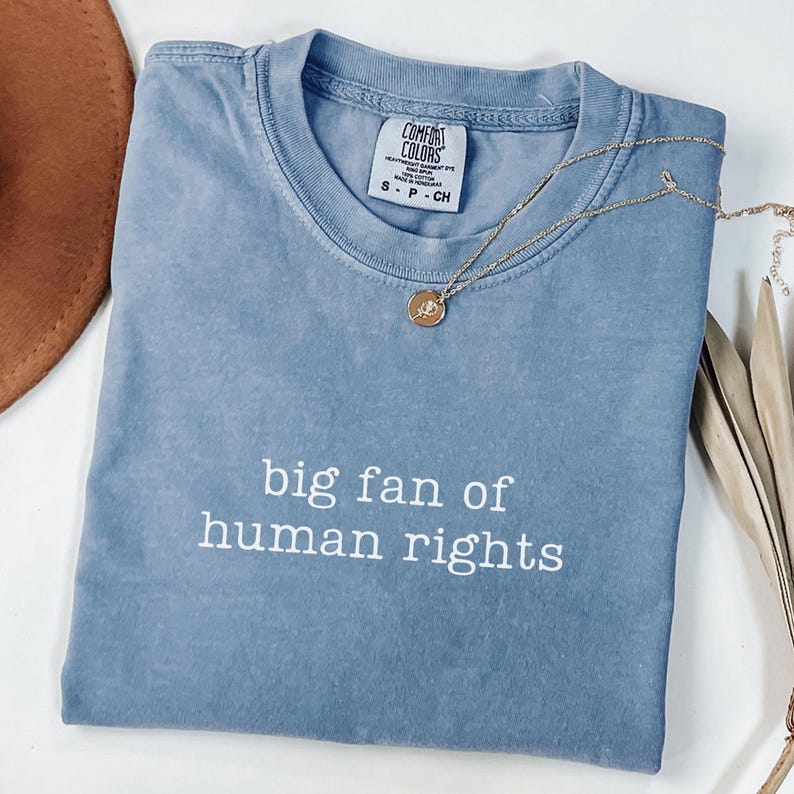 May include: A blue Comfort Colors t-shirt with the text "big fan of human rights" in white. A gold necklace with a pendant rests on the shirt. The shirt is folded and has a tag.
