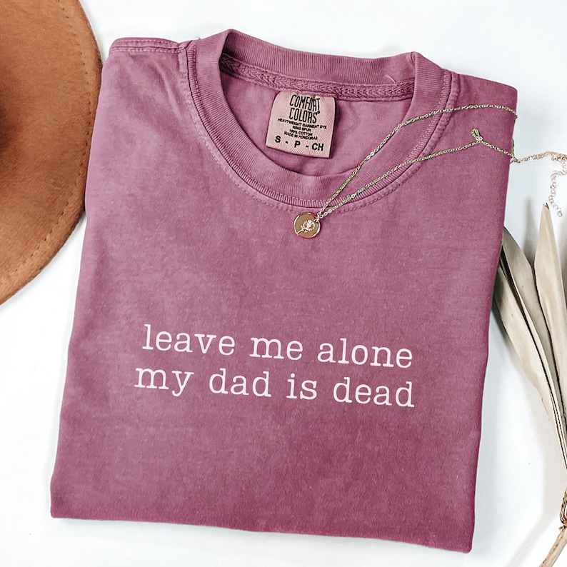 Peut inclure: Un t-shirt Comfort Colors rose poussi&eacute;reux avec le texte "leave me alone my dad is dead" en blanc. Un collier dor&eacute; avec un pendentif est drap&eacute; sur le t-shirt. Un chapeau marron est visible en haut &agrave; gauche.