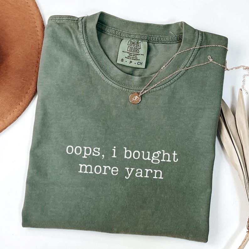 May include: An olive green t-shirt with the text "oops, i bought more yarn" in white. A gold necklace with a pendant is draped on the shirt. The shirt is folded and has a tag that says "Comfort Colors".