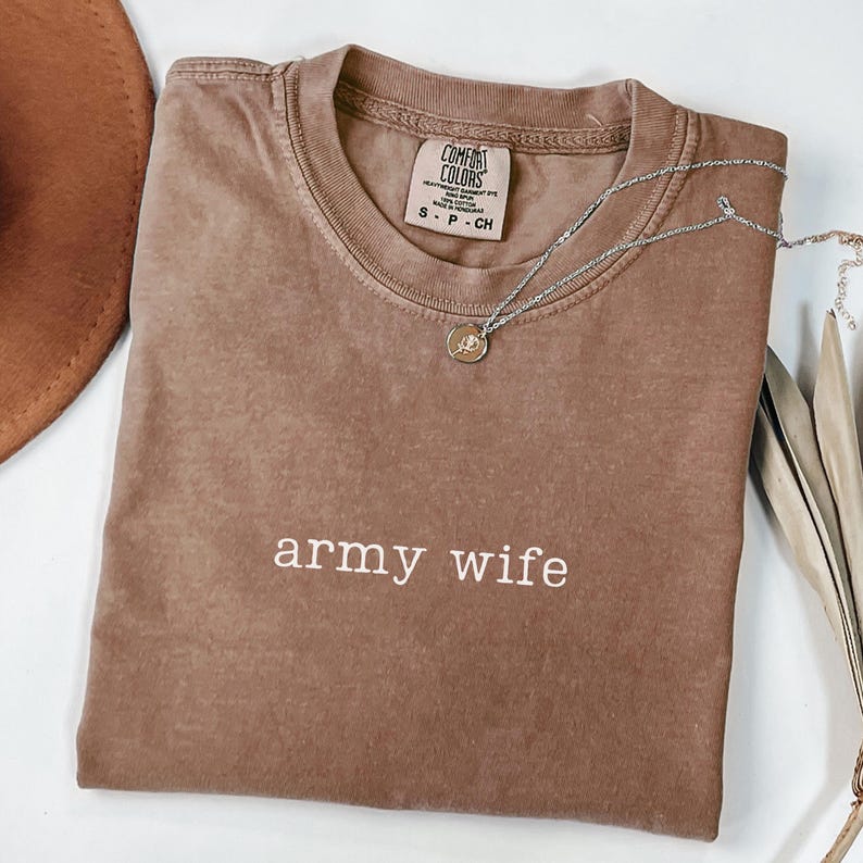 May include: A brown Comfort Colors t-shirt with the words "army wife" in white. A silver necklace with a small pendant rests on the shirt. A brown hat and decorative leaves are in the background.