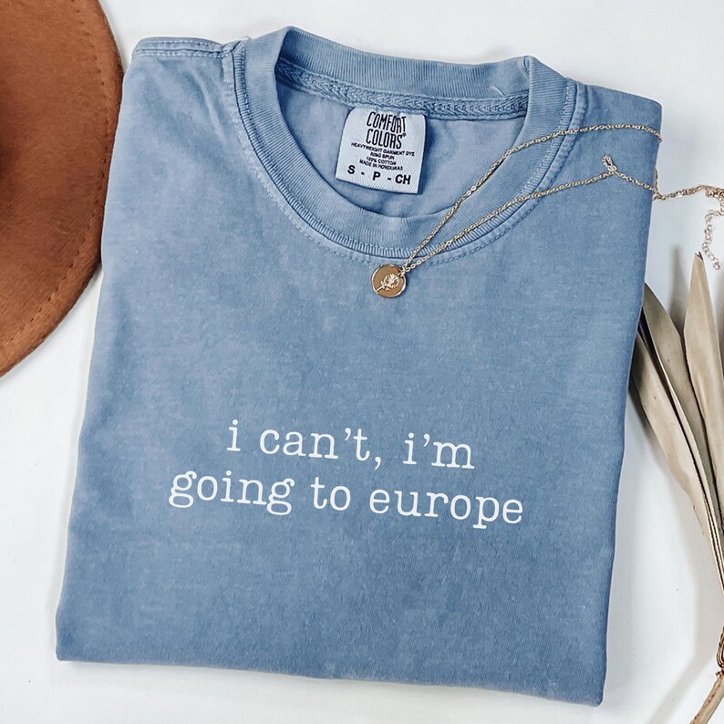 May include: A blue short-sleeved t-shirt with a crew neckline and a Comfort Colors brand label at the collar. The front features the white text "i can't, i'm going to europe" in a simple font. A gold-toned necklace with a small round pendant rests on top.