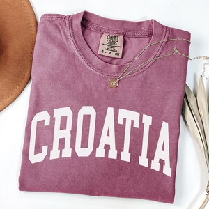 May include: A mauve short-sleeved t-shirt with the word CROATIA printed in large white capital letters across the chest. The shirt features a Comfort Colors brand label at the inner neckline, indicating size S, and is made of 100% cotton.