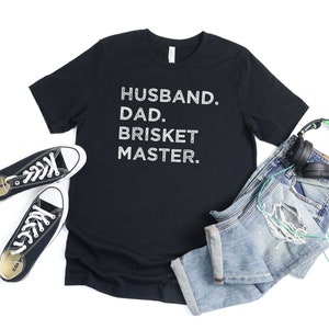 May include: Black t-shirt with white text that reads "Husband. Dad. Brisket Master."