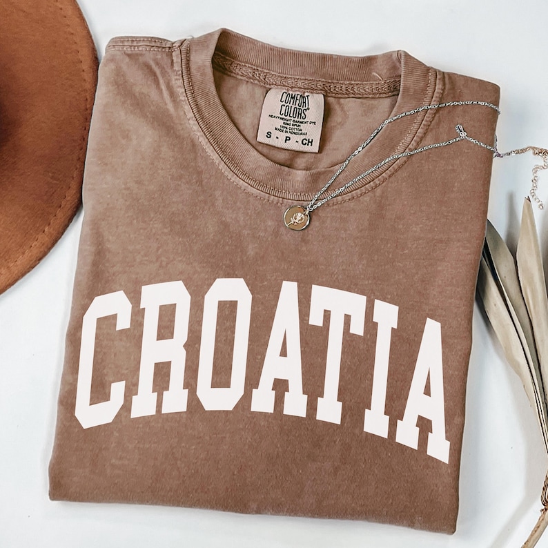 May include: A brown short-sleeved t-shirt with a vintage-wash finish featuring the word CROATIA printed in large white block letters across the chest. The shirt has a crew neckline and a Comfort Colors brand label inside the collar.