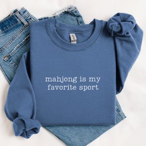 Puede incluir: Una sudadera azul de cuello redondo con el texto bordado en blanco "mahjong is my favorite sport" centrado en el pecho. La prenda tiene mangas largas con pu&ntilde;os acanalados y un cuello acanalado, combinada con unos pantalones vaqueros azules.