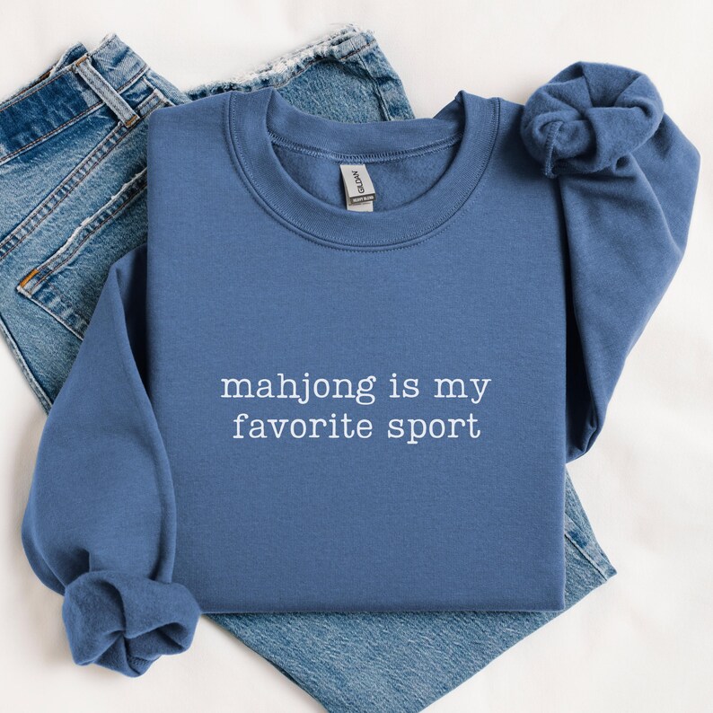 Puede incluir: Una sudadera azul de cuello redondo con el texto bordado en blanco "mahjong is my favorite sport" centrado en el pecho. La prenda tiene mangas largas con pu&ntilde;os acanalados y un cuello acanalado, combinada con unos pantalones vaqueros azules.