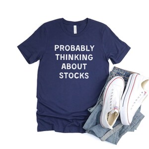 Puede incluir: Una camiseta azul marino con el texto "PROBABLY THINKING ABOUT STOCKS" en letras blancas.