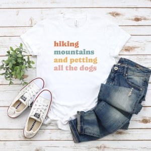 May include: White t-shirt with the text "hiking mountains and petting all the dogs" in colorful letters. The shirt is laid out on a wooden surface with a pair of white sneakers and blue jeans.