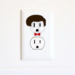 May include: A white electrical outlet with a sticker of a person's face with brown hair and a red bow tie. The person's face is a stylized version of an electrical outlet.