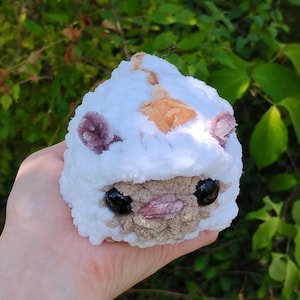 May include: A handmade plush toy in the shape of a round creature. The toy is primarily white with accents of tan, purple, and pink. It has large black eyes and is held in a hand against a backdrop of green foliage.