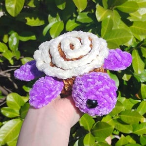 Peut inclure: Une tortue en peluche faite à la main, avec une carapace en spirale blanche et marron, un corps violet et des yeux noirs. La tortue est faite de fil doux et texturé, tenue dans une main devant des feuilles vertes.