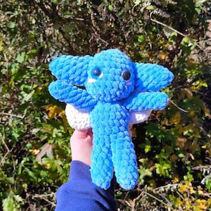 May include: A handmade blue crochet octopus plushie with large eyes and a smiling face. The plushie has a white underside and is held against a backdrop of green foliage. The octopus has multiple tentacles and a textured surface.