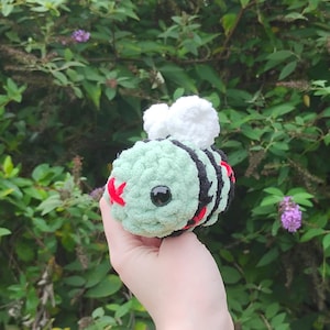 May include: A handmade crochet bee plushie with a light green body, black stripes, and red accents. The bee has large black eyes and white fluffy wings. The plushie is held against a backdrop of green foliage.