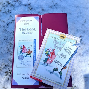 Könnte beinhalten: Eine Nahaufnahme eines Buches mit dem Titel "The Long Winter" von Laura Ingalls Wilder. Das Buch ist auf einer Seite mit einer Illustration von Kindern, die im Schnee spielen, aufgeschlagen. Das Buch liegt auf einem roten Ordner mit einem weißen Lesezeichen.