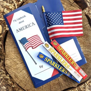 May include: A patriotic lapbook about America, with a small American flag and two boxes of sparklers. The lapbook is open, displaying the title "My Lapbook about AMERICA". The sparklers are red and blue.