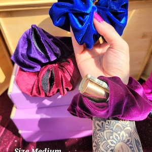 Może przedstawiać: Cztery scrunchies z aksamitu w różnych kolorach: niebieski, fioletowy, bordowy i ciemnofioletowy. Scrunchies są ułożone na fioletowym pudełku z napisem "Rozmiar średni".
