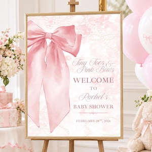 May include: A rectangular sign on a wooden easel featuring a large pink satin bow illustration on the left and text that reads Tiny Toes & Pink Bows WELCOME TO Rachel's BABY SHOWER FEBRUARY 28TH, 2026 against a soft pink watercolor background.