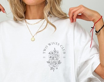 May include: White t-shirt with a black graphic design that says "Two Wives Club" with a floral design in the center.