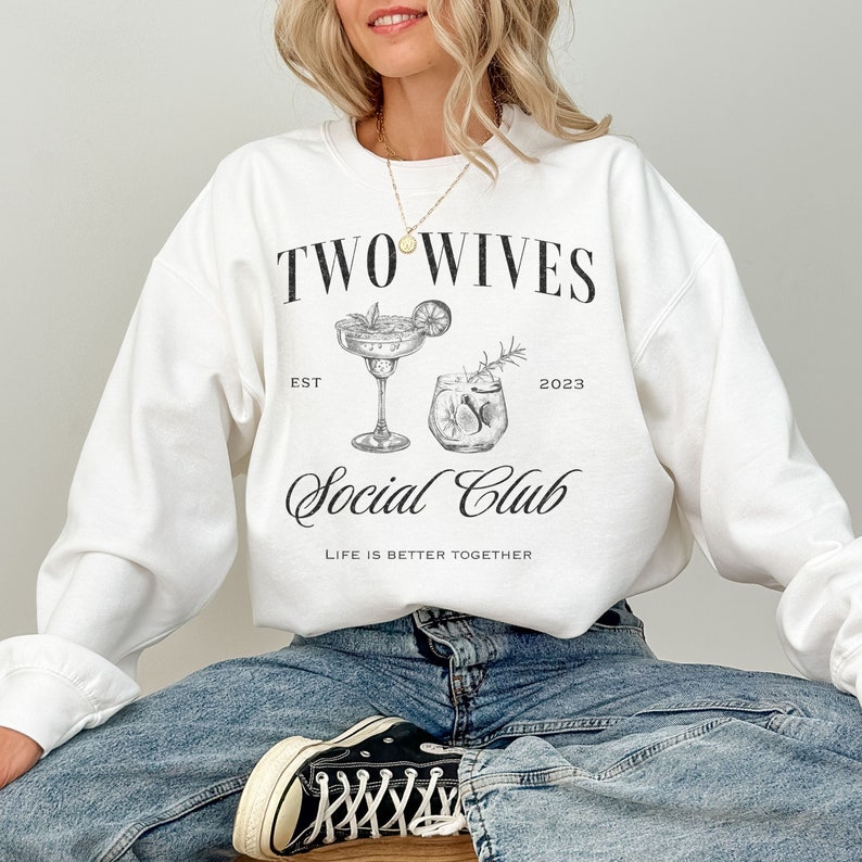 Puede incluir: Sudadera blanca con el texto "TWO WIVES SOCIAL CLUB" e ilustraciones de c&oacute;cteles. La sudadera tambi&eacute;n incluye el texto "EST 2023" y "LIFE IS BETTER TOGETHER".