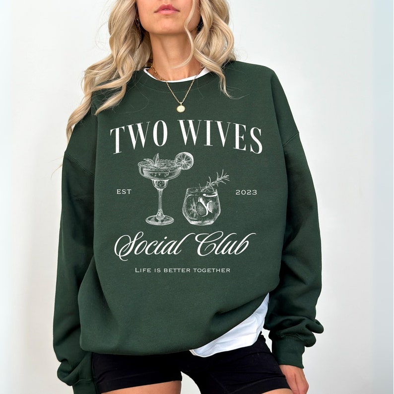 Puede incluir: Una sudadera verde oscuro con el texto "TWO WIVES Social Club" en blanco. El dise&ntilde;o incluye ilustraciones de dos c&oacute;cteles y la frase "LIFE IS BETTER TOGETHER".