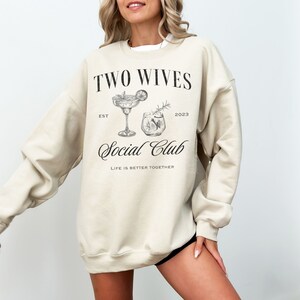 Puede incluir: Sudadera beige con el texto "TWO WIVES Social Club" en escritura negra. El dise&ntilde;o incluye ilustraciones de dos c&oacute;cteles, con el texto "EST 2023" y "LIFE IS BETTER TOGETHER". Una prenda informal y c&oacute;moda.