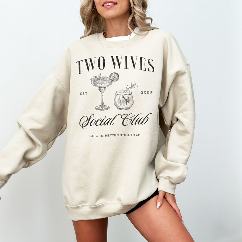 Puede incluir: Sudadera beige con el texto "TWO WIVES Social Club" en escritura negra. El dise&ntilde;o incluye ilustraciones de dos c&oacute;cteles, con el texto "EST 2023" y "LIFE IS BETTER TOGETHER". Una prenda informal y c&oacute;moda.